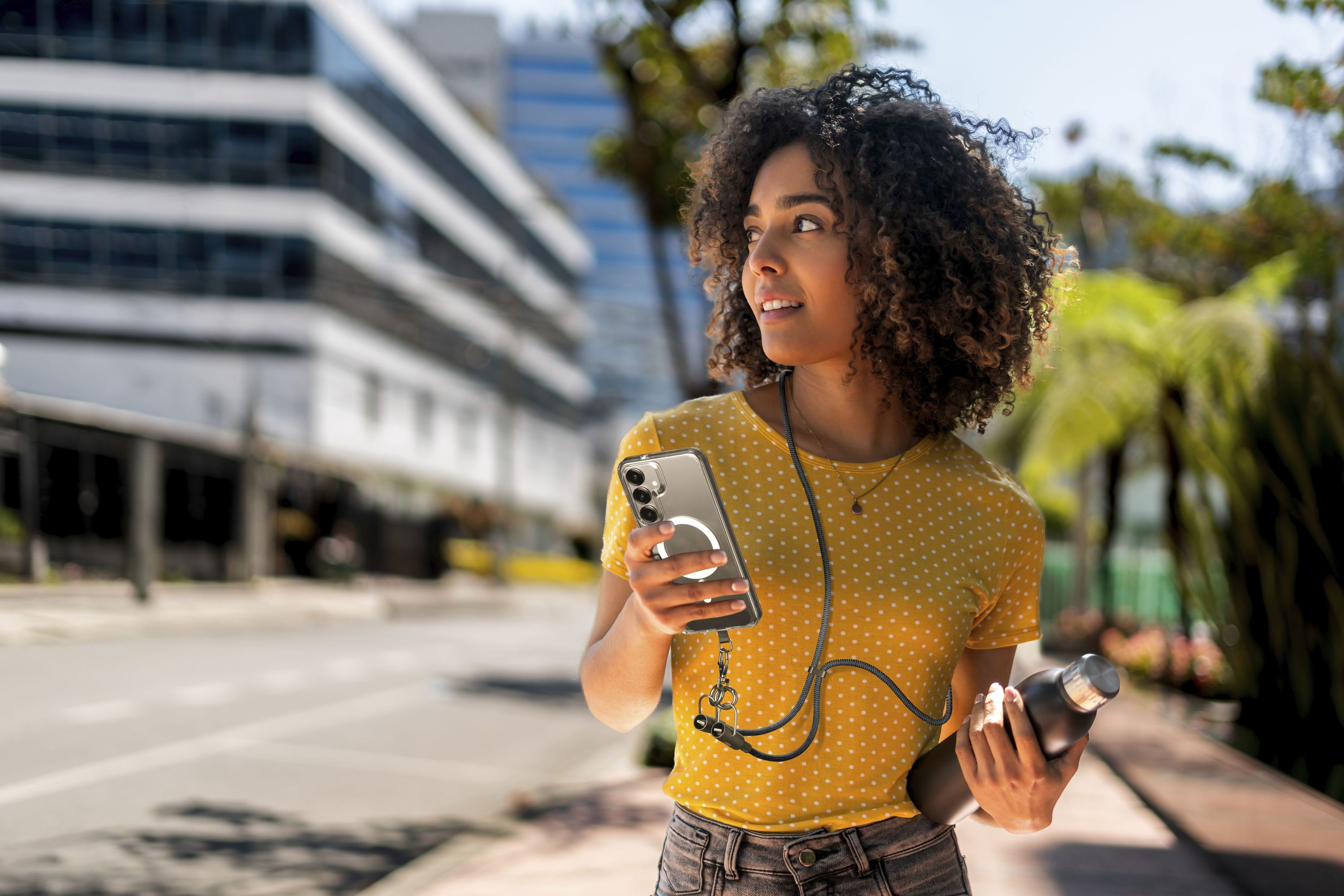 Kıvırcık saçlı bir kadın dışarıda telefon ve su şişesi tutuyor.