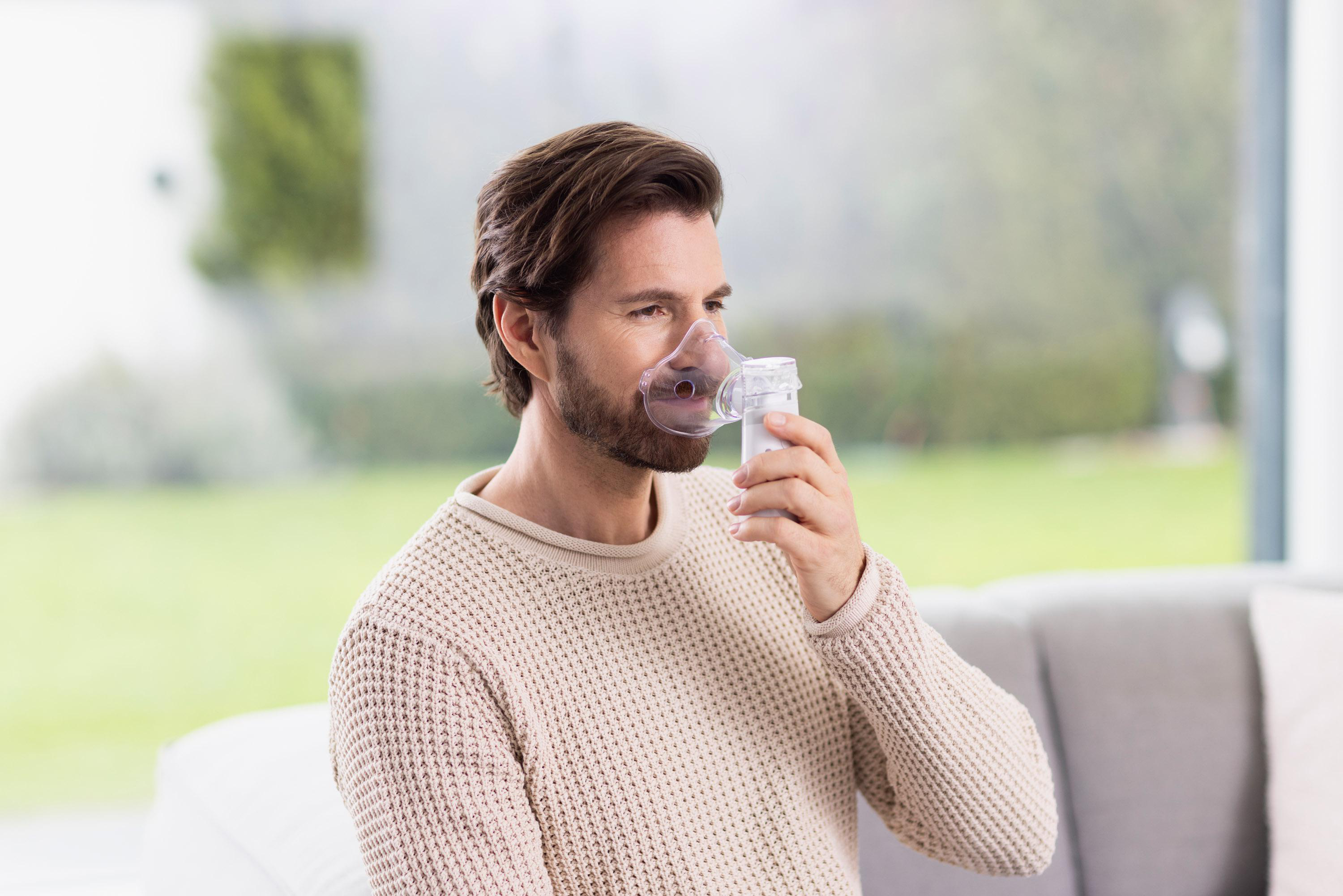Ein Mann mit Bart trägt eine Inhalatormaske und hält das Gerät in der Hand.