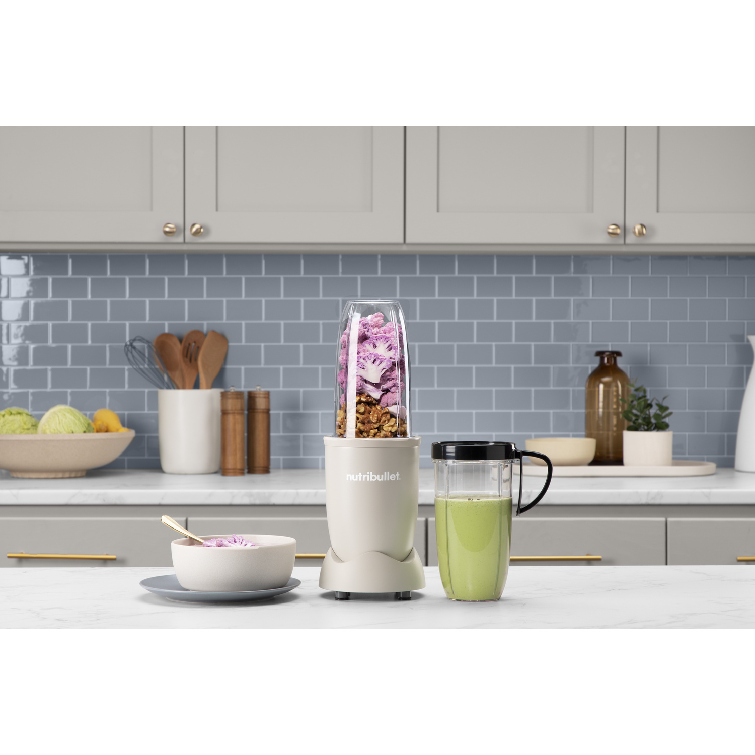 Un frullatore NutriBullet beige con fiori viola, noci e una bevanda verde su un piano di lavoro in marmo.