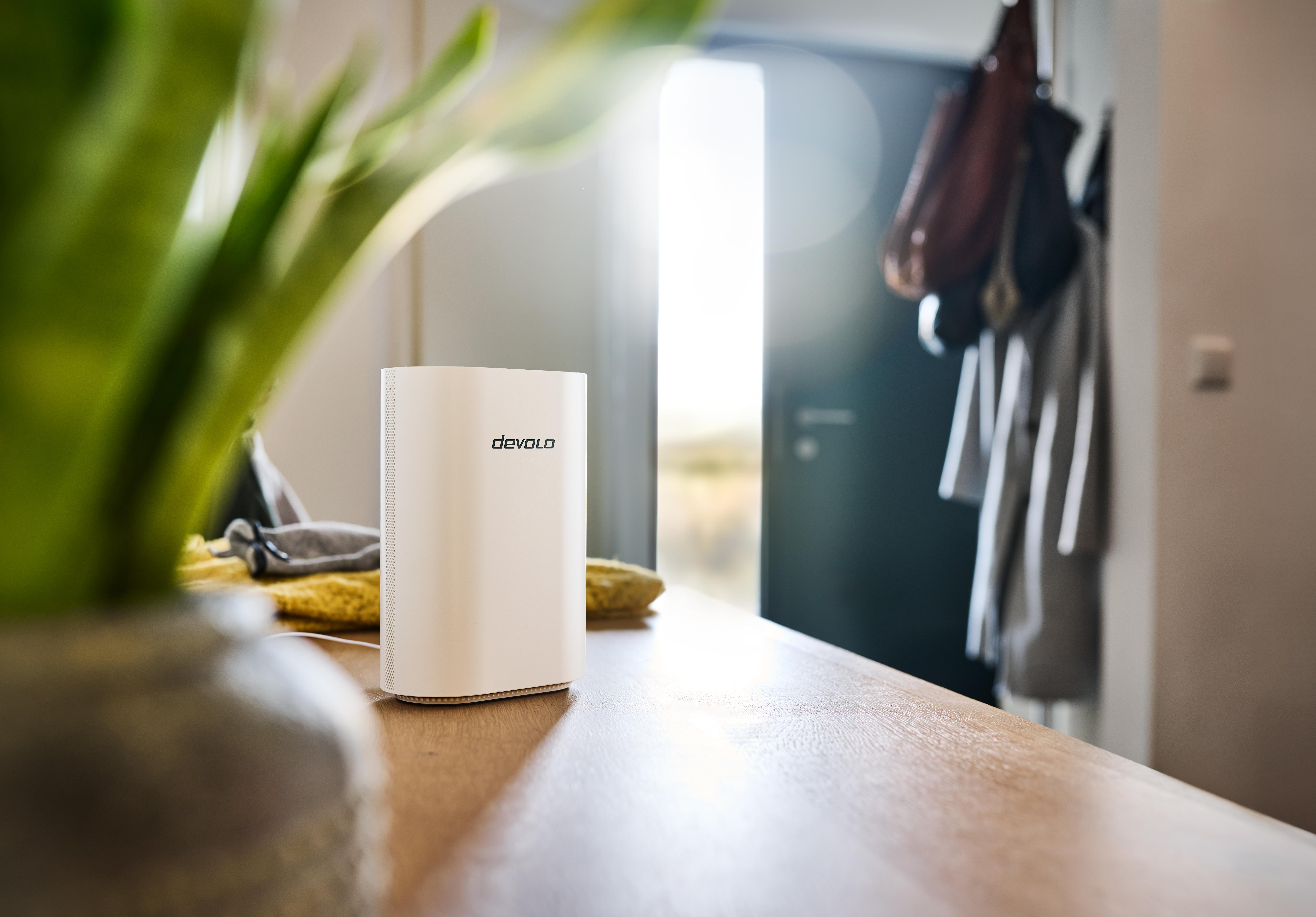 Ein weißer DEV-OLO-Router steht auf einem Holztisch, im Hintergrund eine Pflanze und eine Tür.