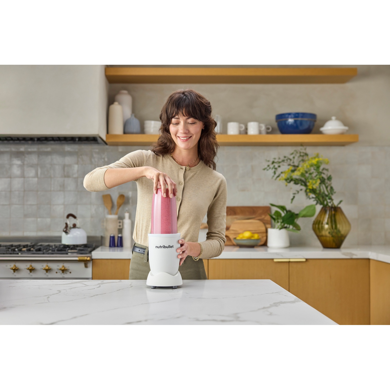 Una donna con maglione beige usa un frullatore Nutribullet bianco con frullato rosa.