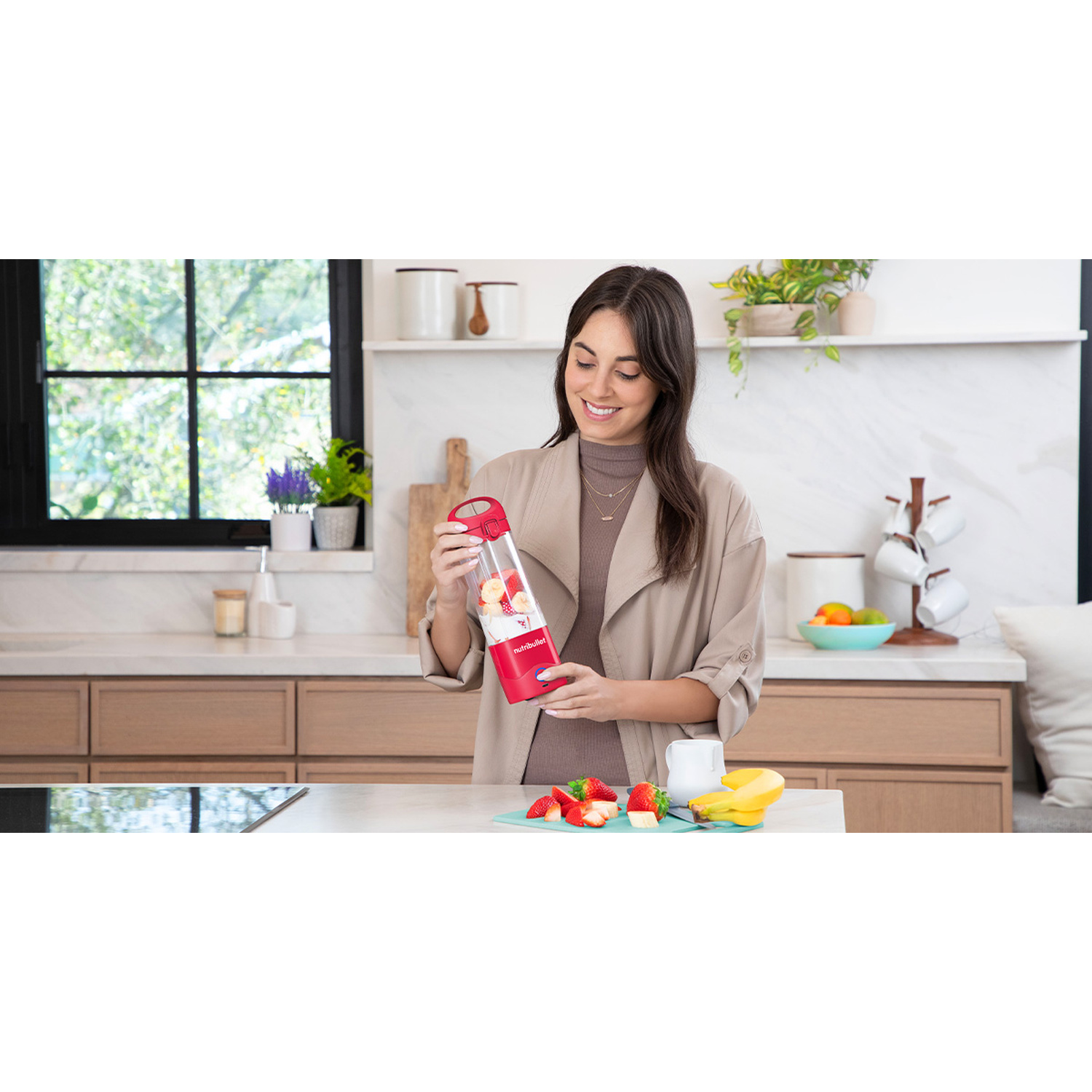 Una donna tiene un frullatore rosso con frutta. Fragole e banane su un tagliere.