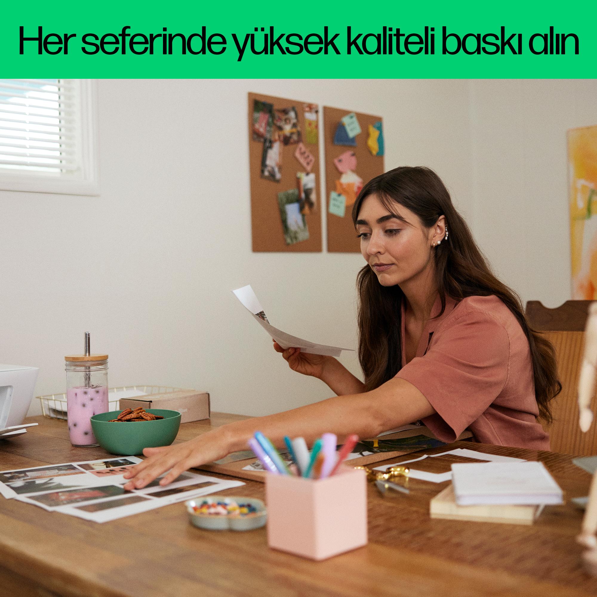 Bir kadın ahşap masada oturuyor, elinde bir kağıt tutuyor. Masada kalemler ve bir vazo var.