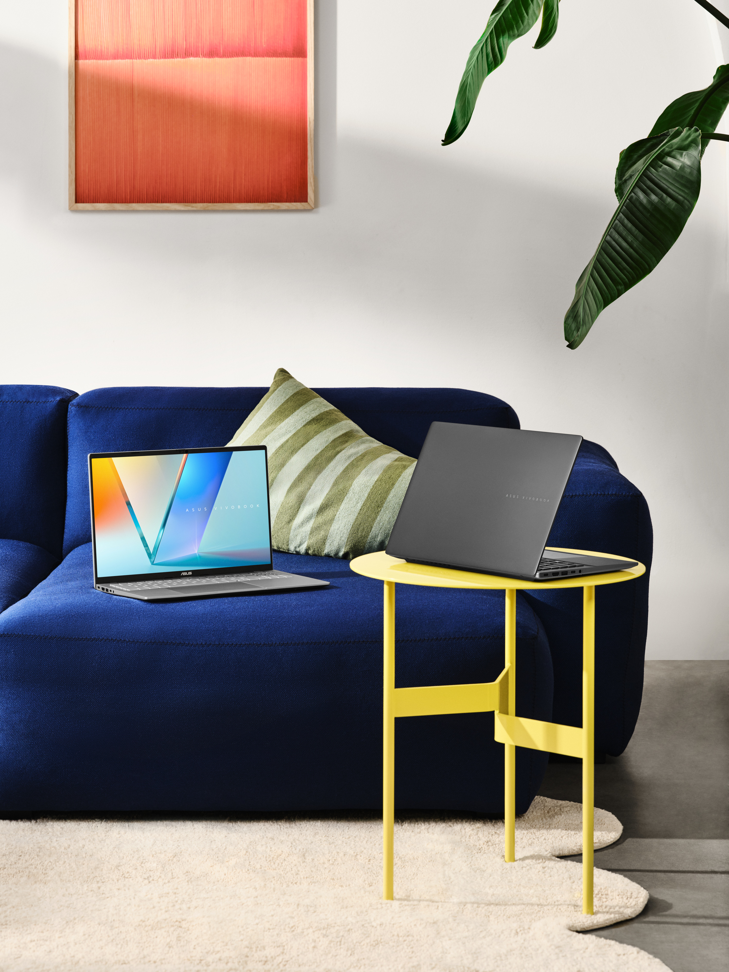 Twee laptops staan op een blauwe bank en een gele bijzettafel. Een rood abstract schilderij en een groene plant zijn op de achtergrond.