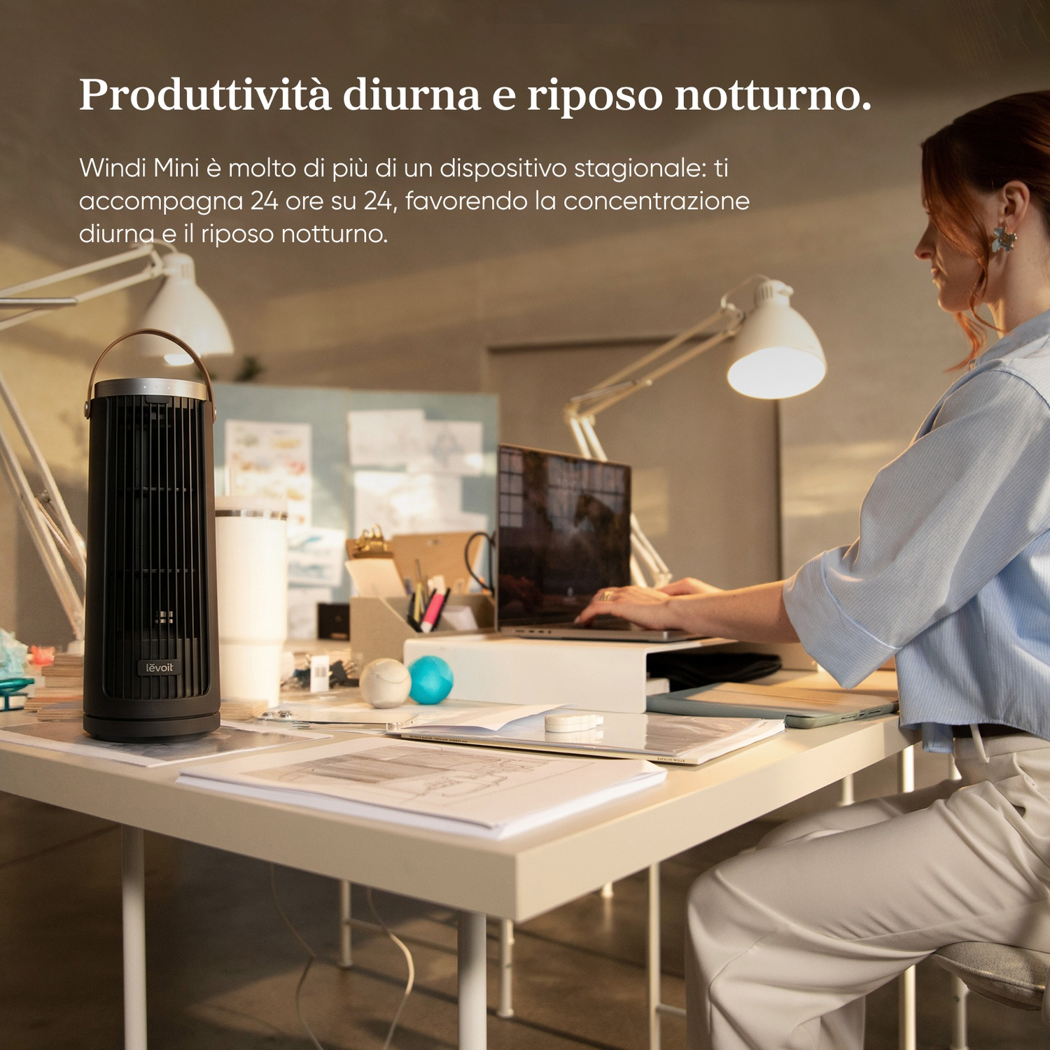 Una donna lavora al computer su una scrivania con ventilatore e carte.