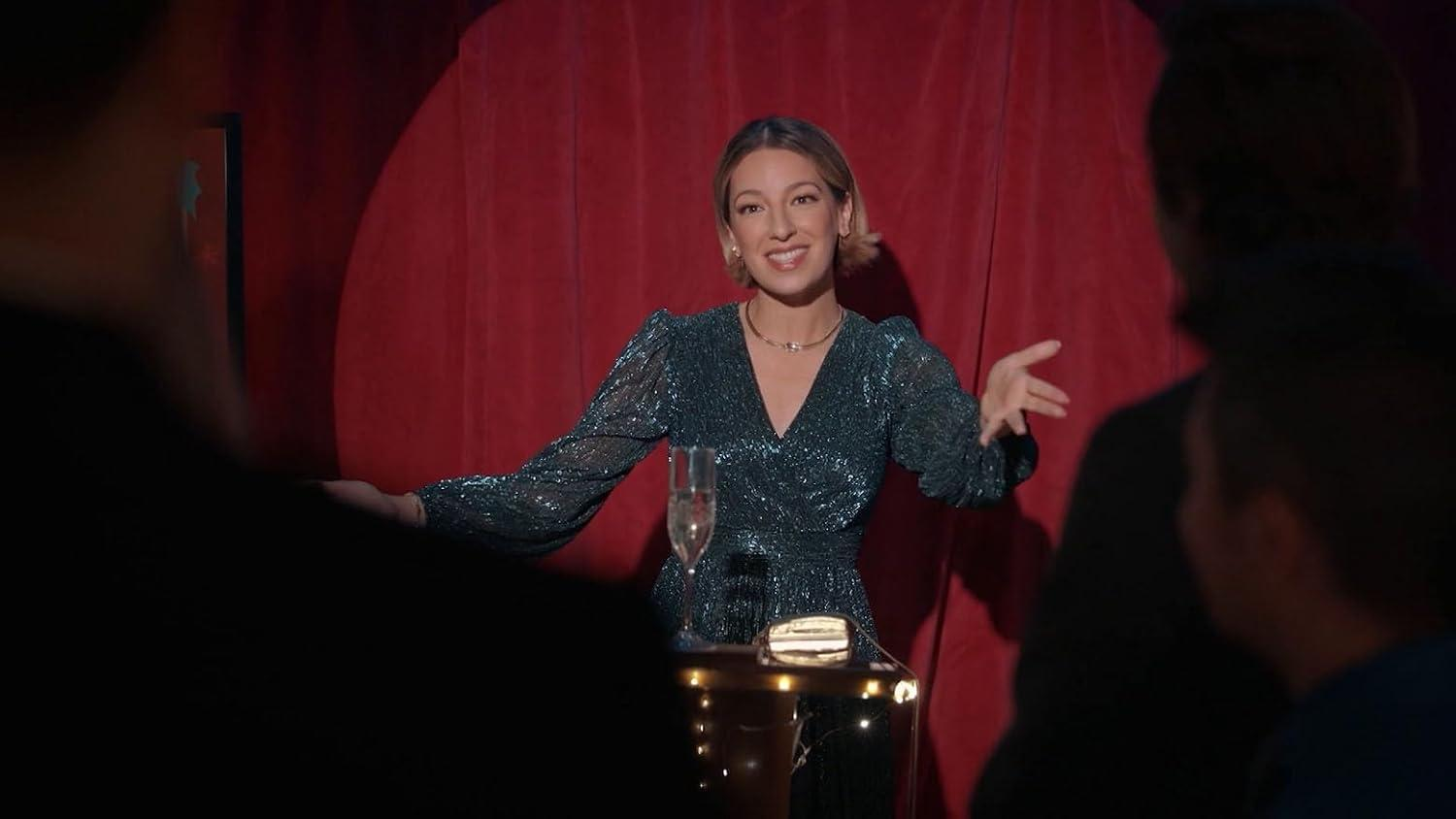 Eine Frau in einem glitzernden Kleid steht vor einem roten Vorhang und spricht.