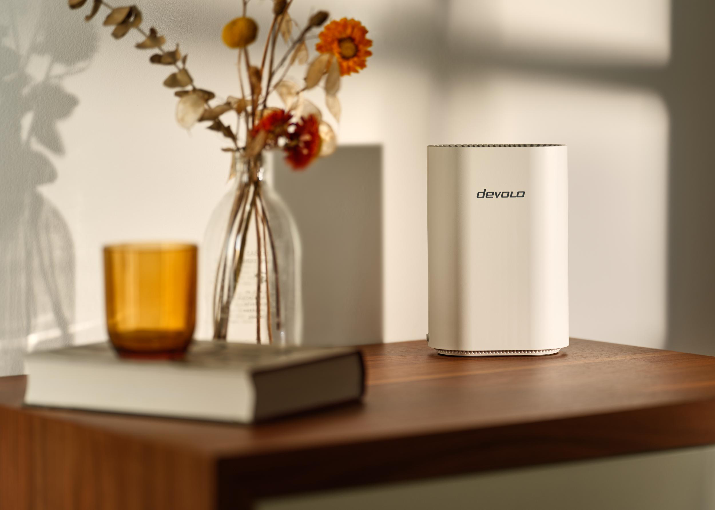 Ein weißer Devolo Router steht auf einem Holztisch neben einem Buch und einer Vase mit Trockenblumen.