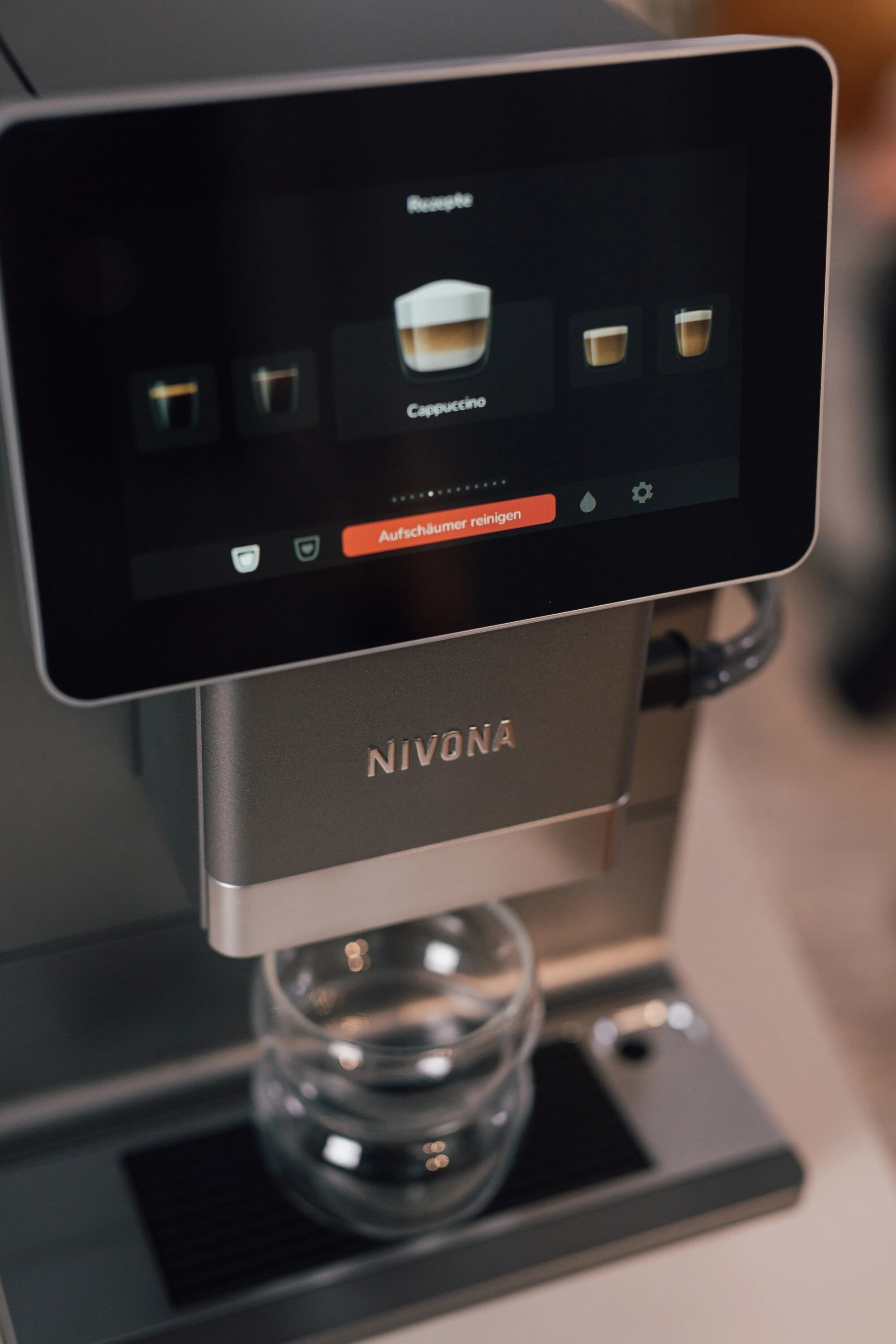 Ekspres do kawy Nivona z ekranem dotykowym wyświetlającym cappuccino. Poniżej stoi filiżanka.
