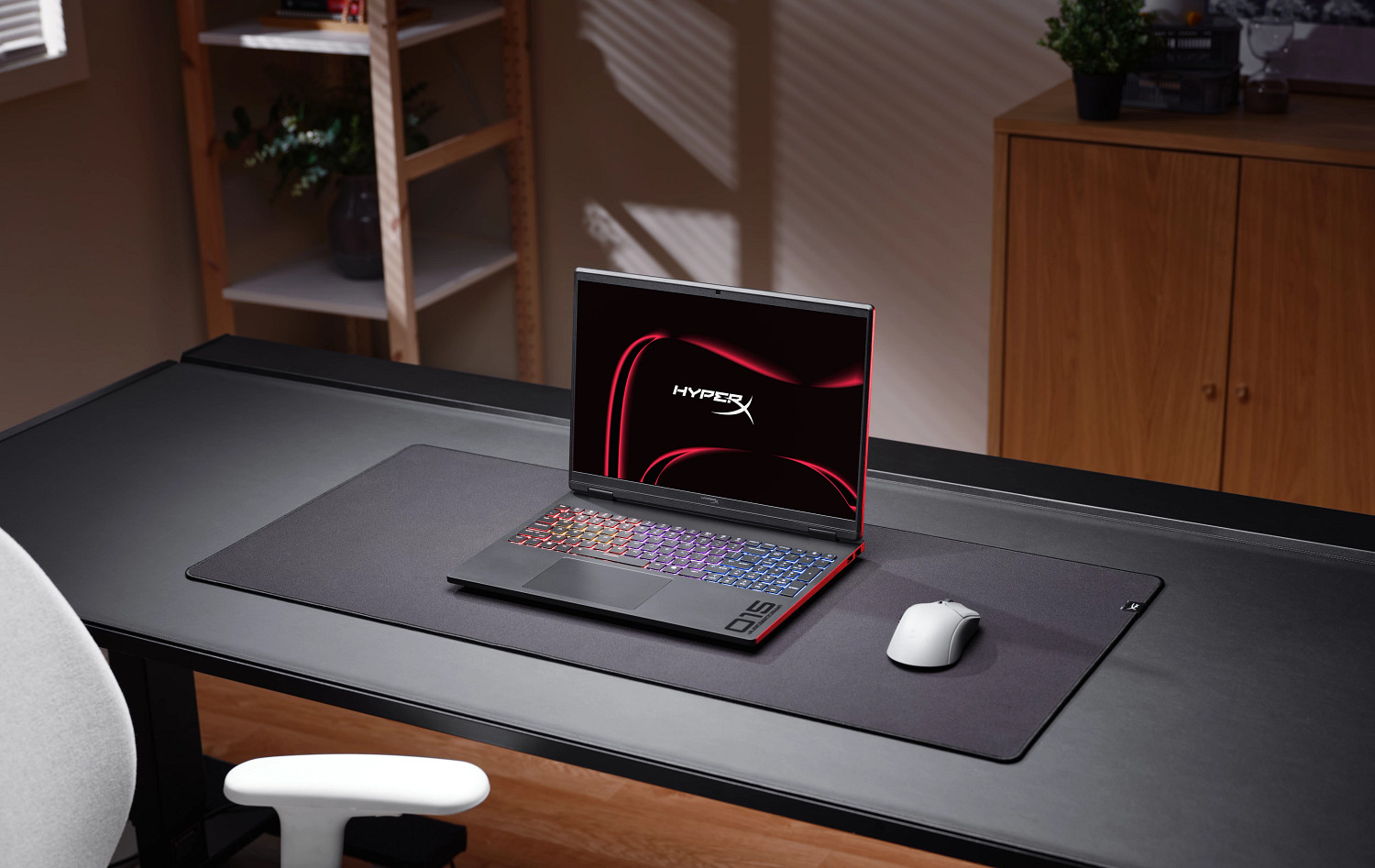 Laptop z czerwonym logo HyperX jest otwarty na biurku z myszą i klawiaturą.