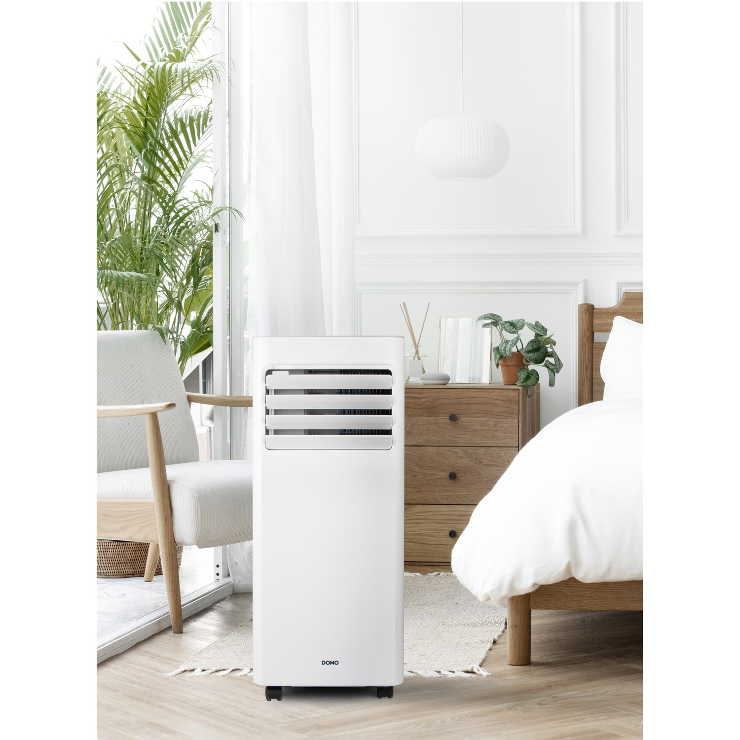 Witte draagbare airconditioner in een lichte slaapkamer met bed, stoel en planten.