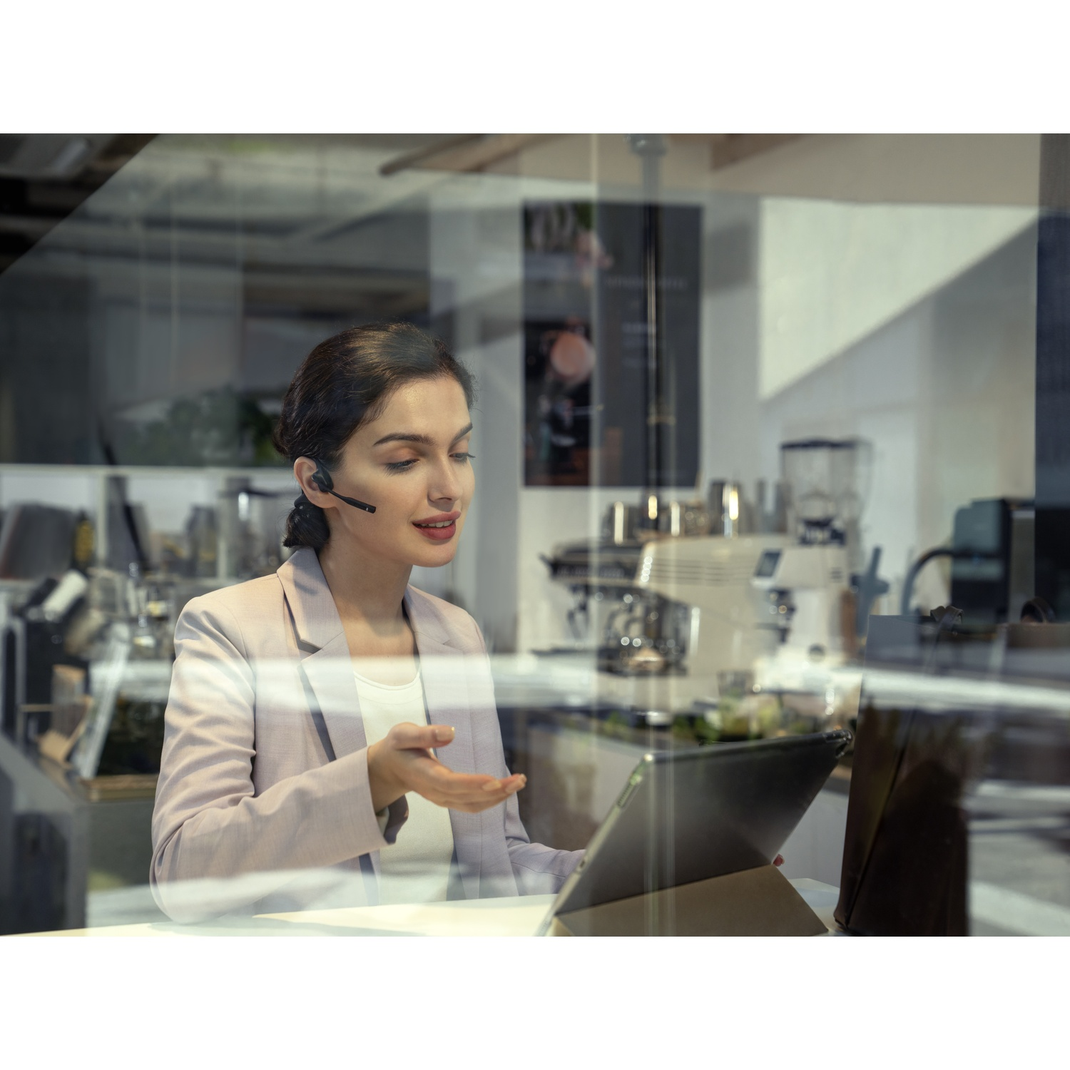 Vrouw met headset praat, gebaart naar tablet in café met espressomachine.