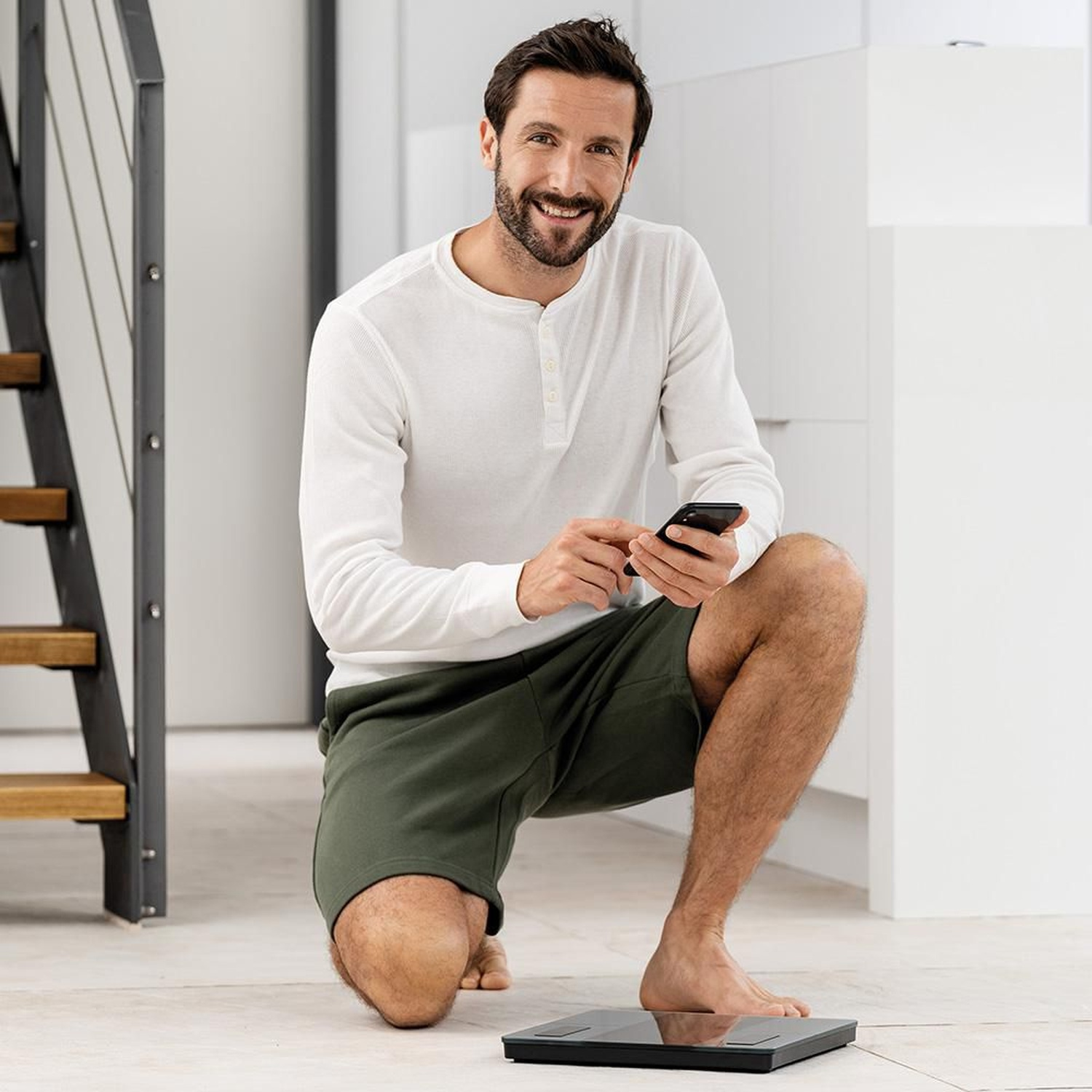 Un uomo in ginocchio su una bilancia tiene uno smartphone. Indossa un top bianco e pantaloncini verdi.