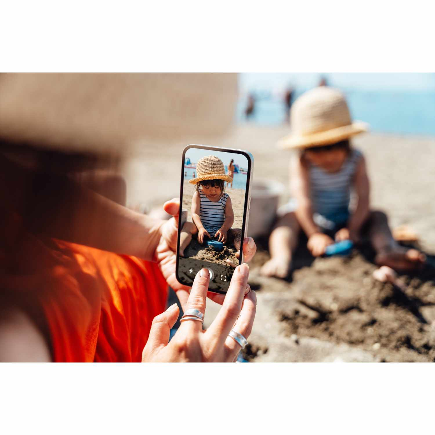 Persona sostiene teléfono capturando niño en sombrero paja jugando en arena playa.