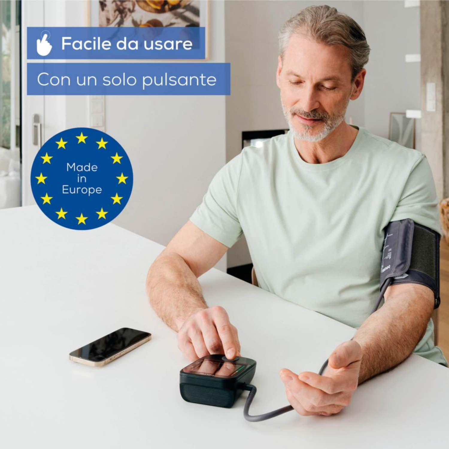 Uomo misura pressione con dispositivo, smartphone e bandiera UE sul tavolo.