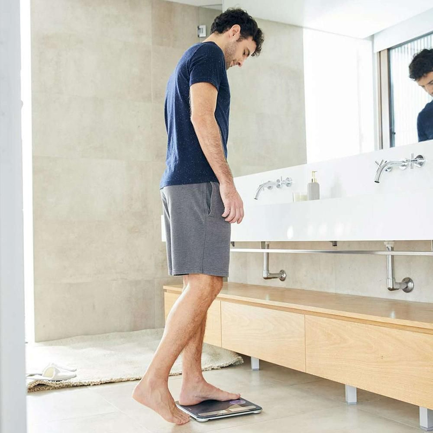Un uomo in maglietta scura e pantaloncini grigi è in piedi su una bilancia in bagno.