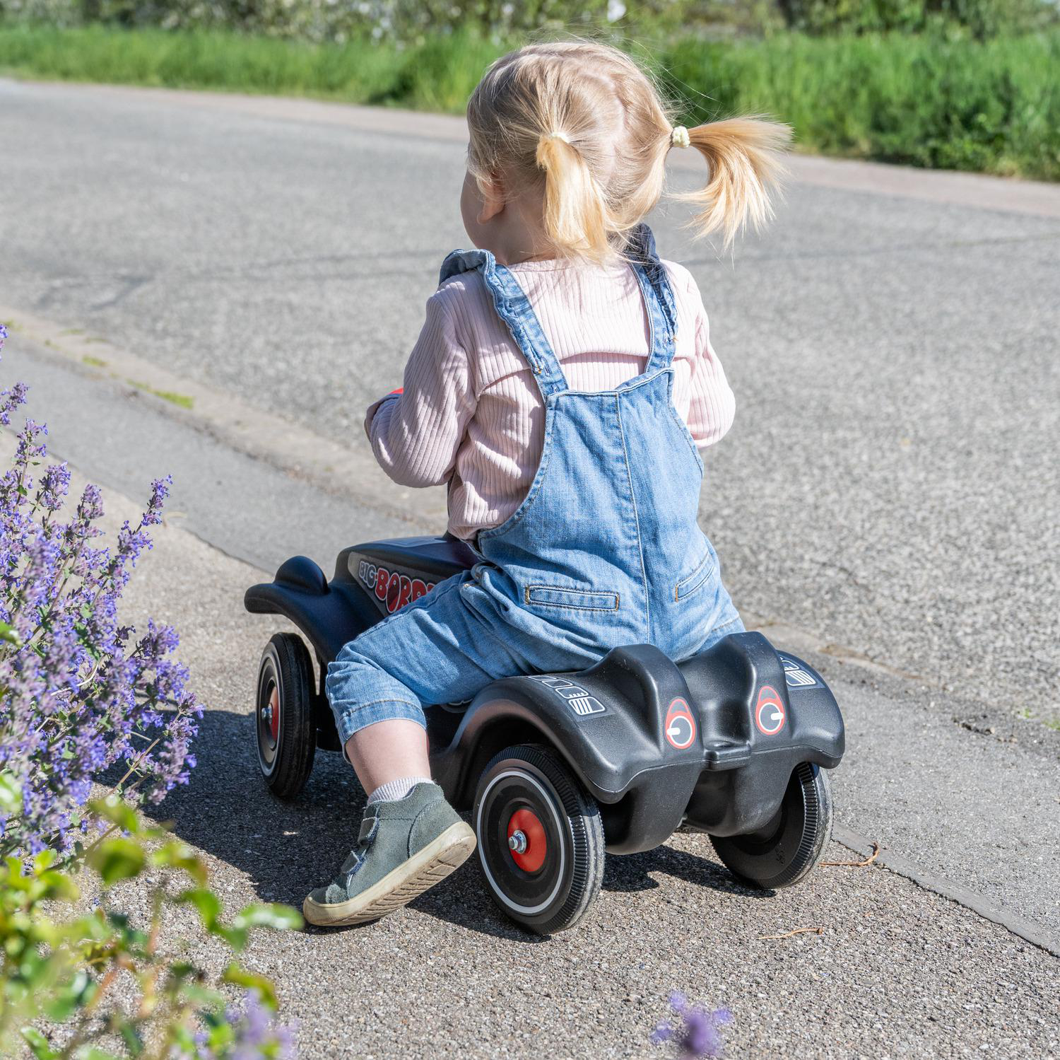 Ein kleines Mädchen in Latzhose fährt auf einem schwarzen Spielzeugauto auf einer Straße.