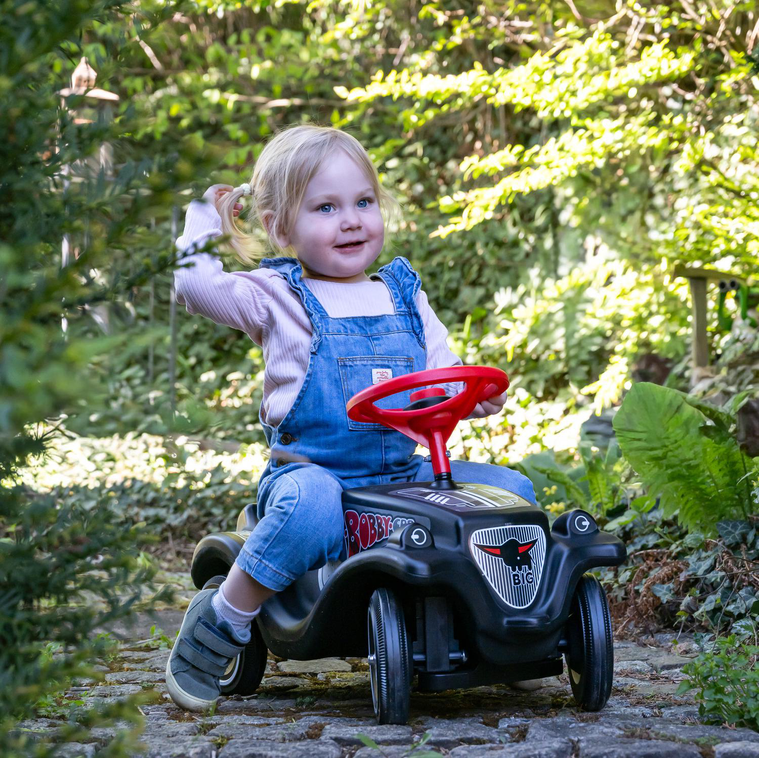 Ein kleines Mädchen mit blauen Augen fährt ein schwarzes Bobby-Car mit rotem Lenkrad im Garten.