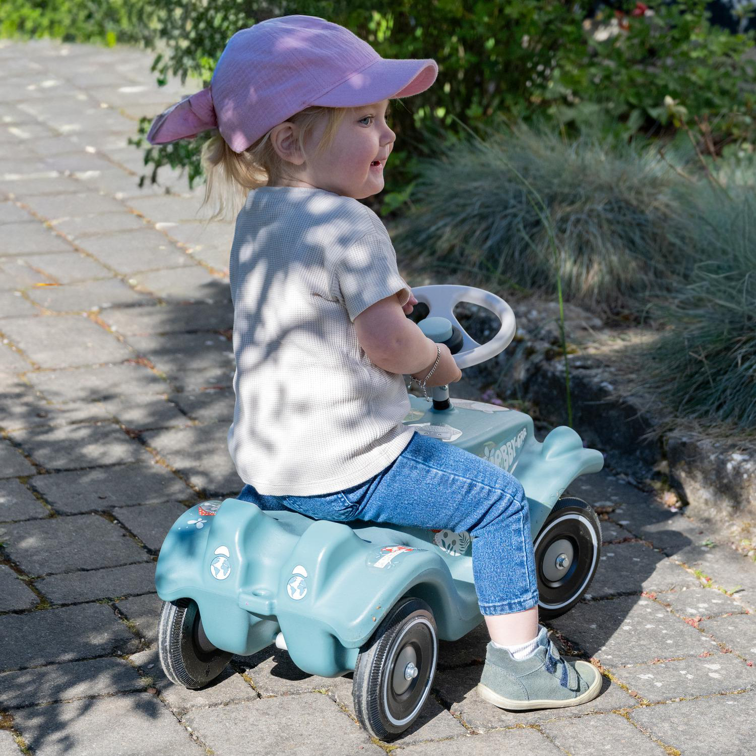 Ein Kleinkind mit rosa Hut sitzt auf einem blauen Spielzeugauto auf einem gepflasterten Weg.