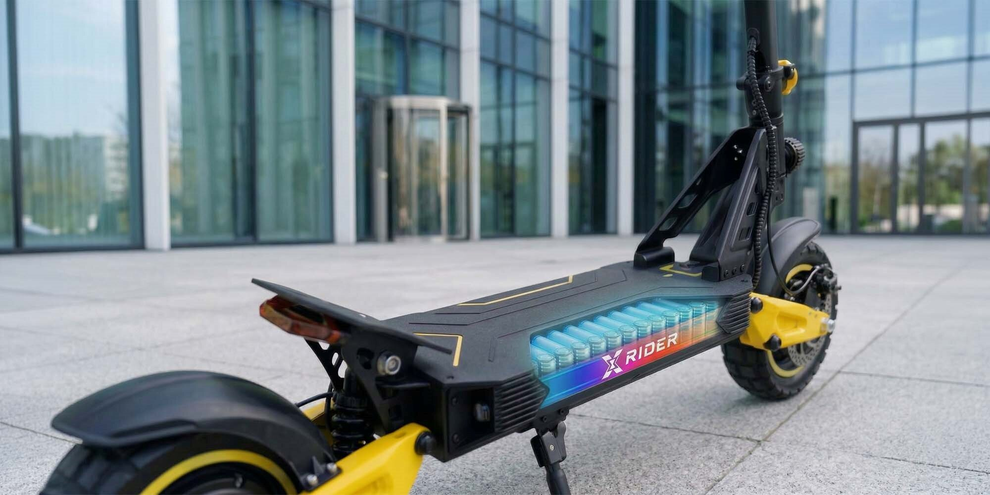 Hulajnoga elektryczna z widoczną baterią i żółtymi akcentami przed nowoczesnym budynkiem.