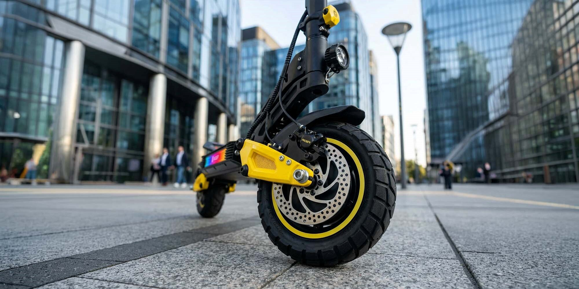 Żółty skuter elektryczny z terenowymi oponami stoi na chodniku przed nowoczesnymi budynkami.