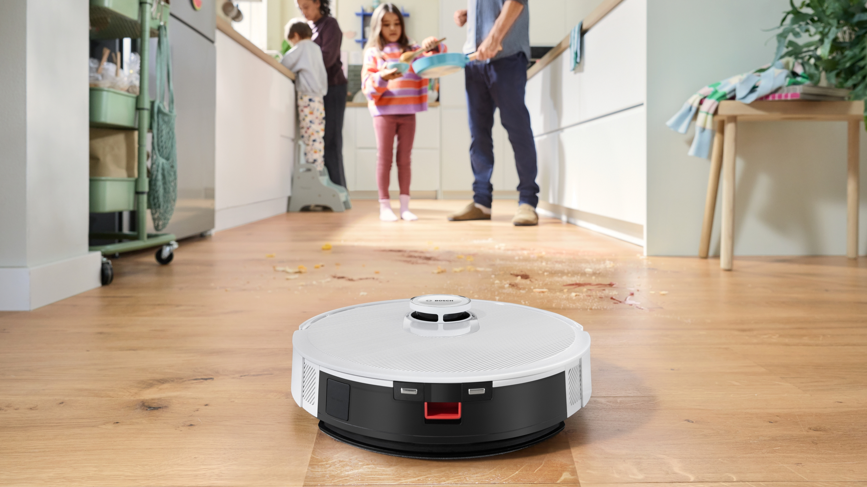 Egy robotporszívó ételmaradékokkal tisztít egy fapadlót, miközben egy család főz.