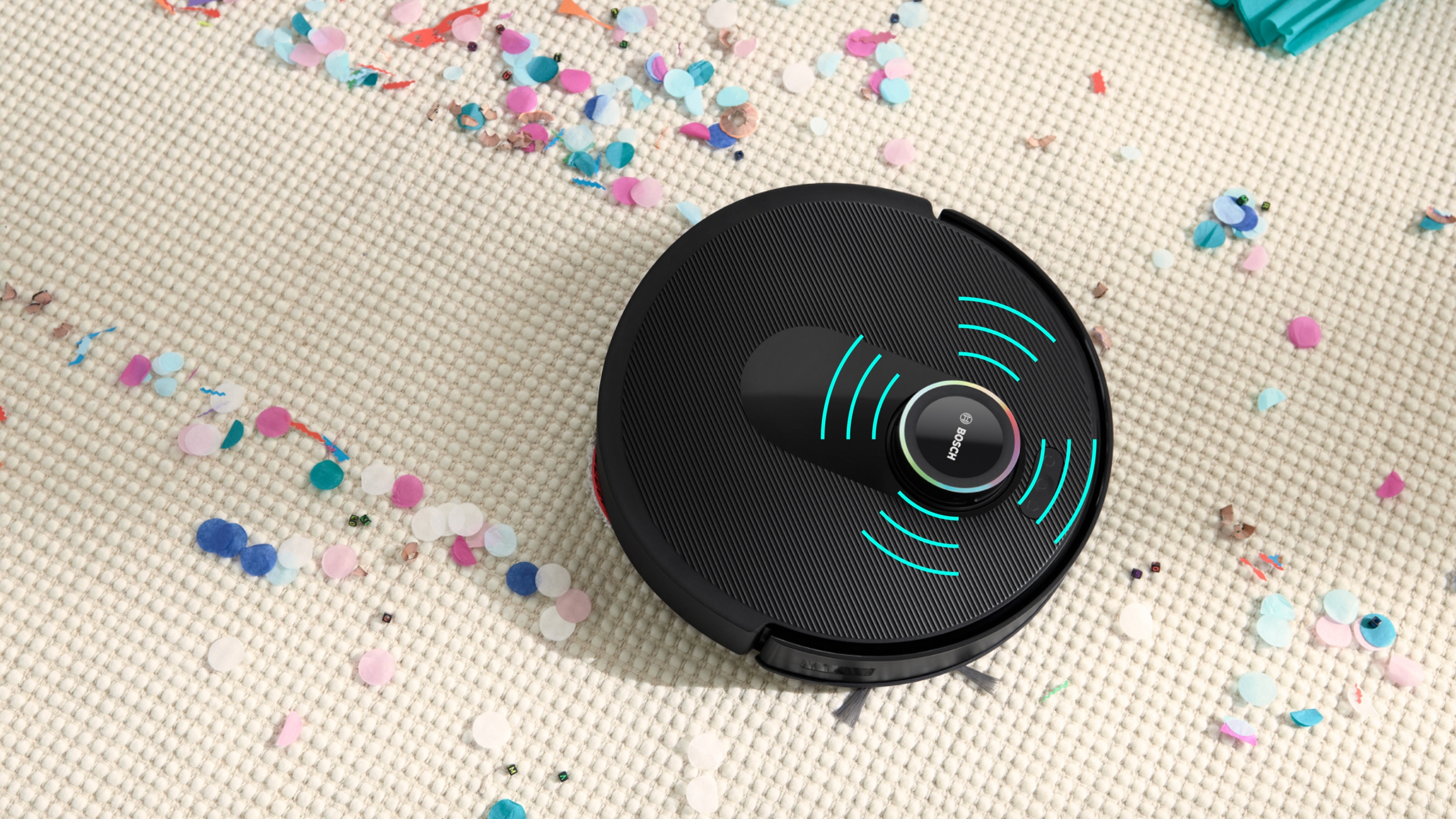Egy fekete robotporszívó színes konfettit tisztít egy texturált krémszínű szőnyegről.