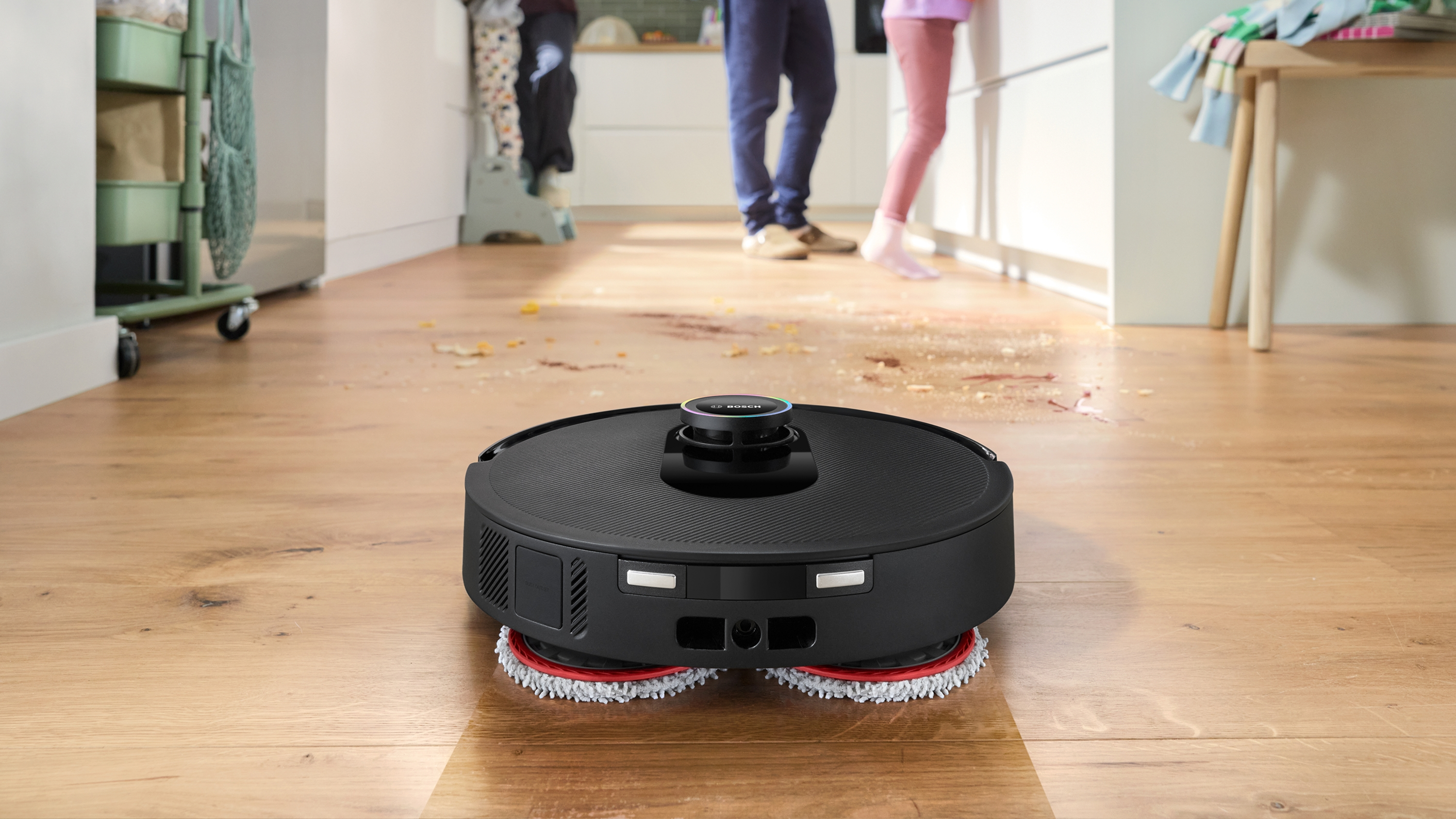 Egy fekete robotporszívó tisztít egy koszos fapadlót, emberekkel a háttérben.