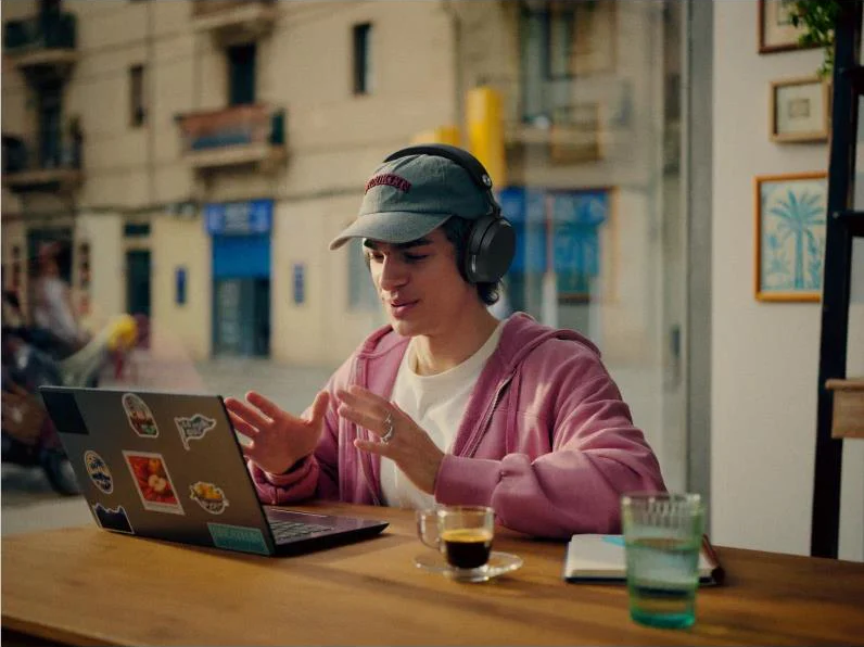 Un uomo con cuffie e felpa con cappuccio è seduto al laptop, beve un caffè.