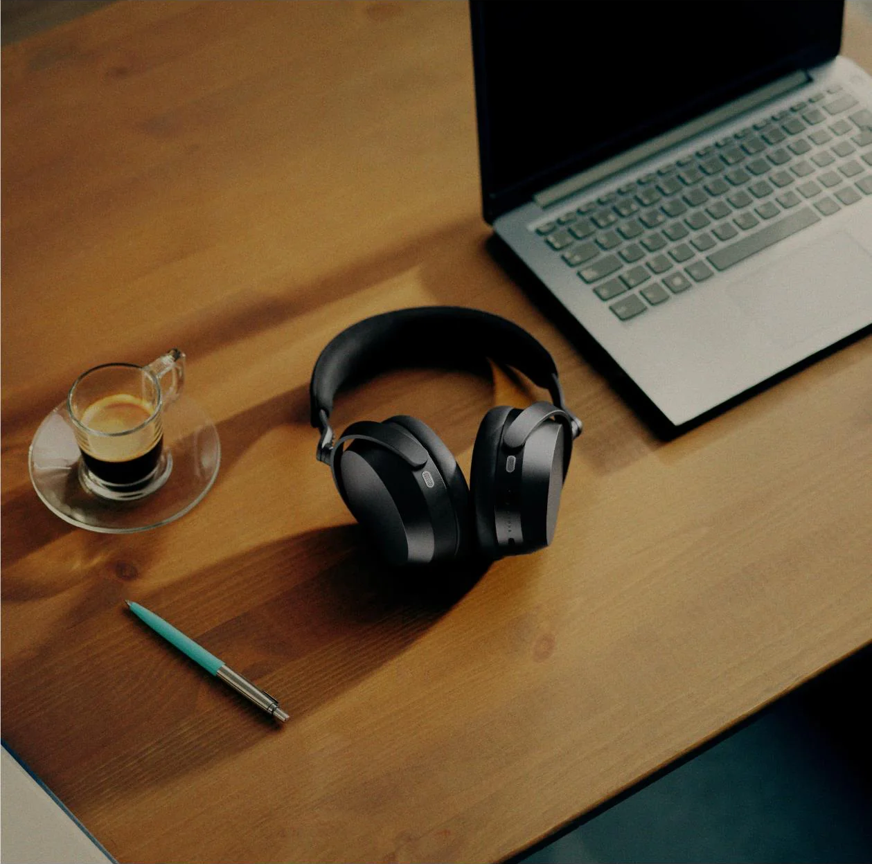 Cuffie nere, caffè, penna e laptop su una scrivania di legno.
