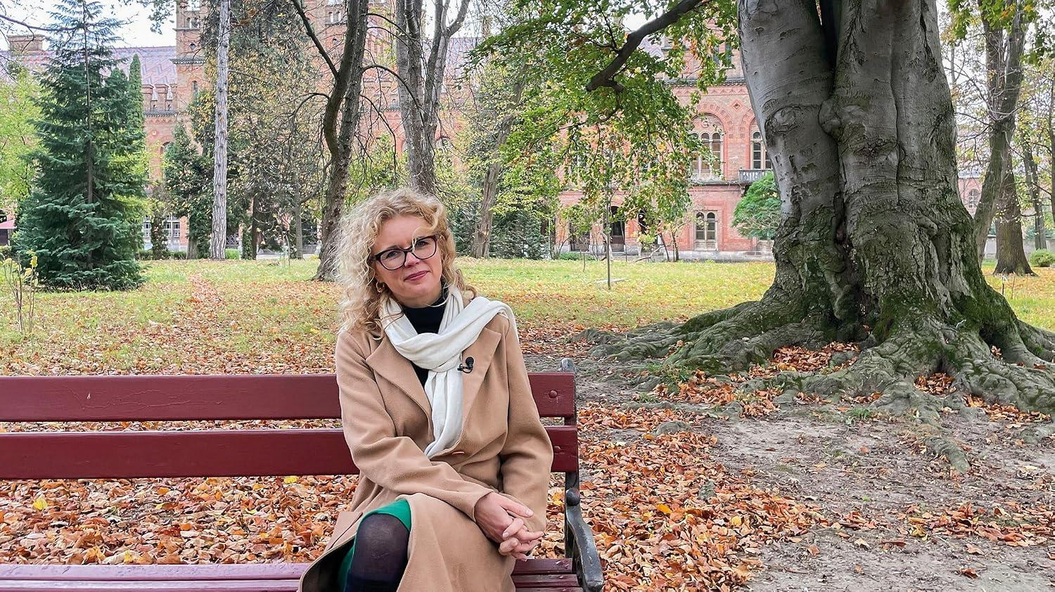 Frau in einem hellbraunen Mantel und Schal sitzt im Herbst auf einer Parkbank, Gebäude und Bäume dahinter.