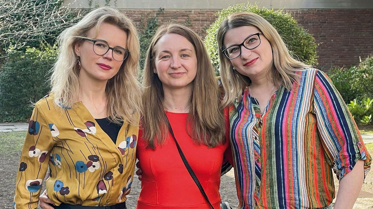 Drei Frauen stehen draußen. Eine trägt ein gelbes Hemd mit Blumen, eine ein rotes Kleid, eine ein gestreiftes Hemd.