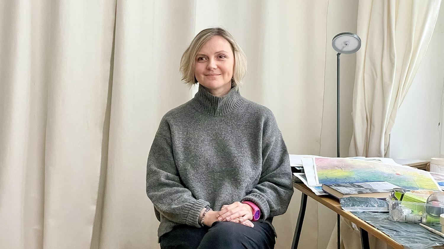 Eine Frau in einem grauen Pullover sitzt vor einer hellen Wand, daneben ein Tisch mit Kunstmaterialien.