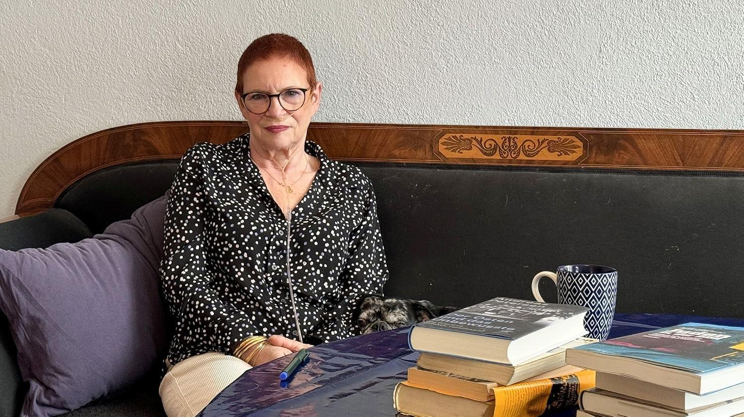 Eine Frau mit roten Haaren sitzt auf einem Sofa neben einem kleinen Hund. Vor ihr liegen Bücher.