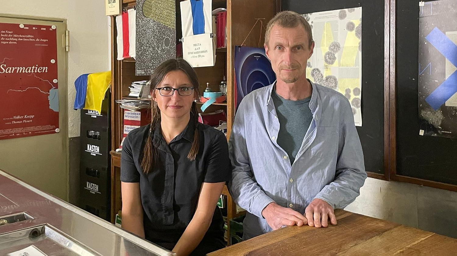 Ein Mann und eine Frau stehen hinter einer Theke. Im Hintergrund sind Regale mit Büchern und Plakaten.
