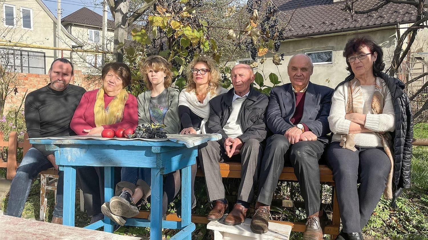Sieben Personen sitzen auf einer Bank und einem Tisch im Freien vor einem Haus.