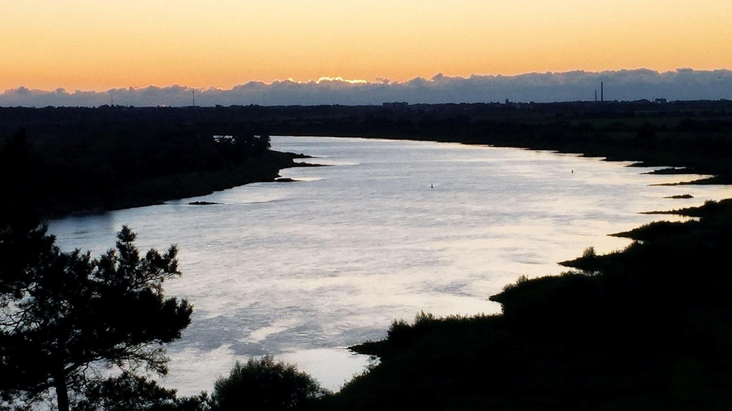Fluss bei Sonnenuntergang mit silhouettierten Bäumen und fernen Wolken.