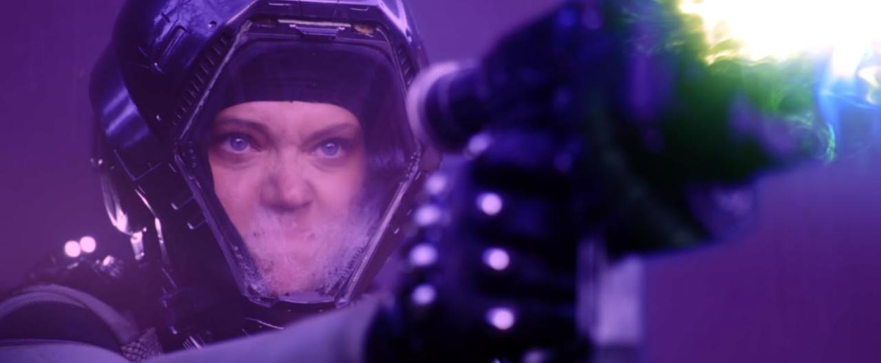 Eine Person in einem futuristischen Helm feuert eine Waffe mit grünem Licht ab.