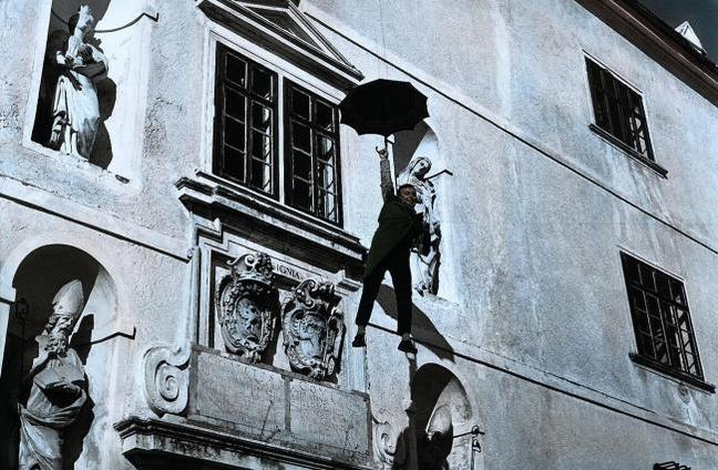 Mann mit Regenschirm hängt an Fassade neben Statuen und Fenstern.