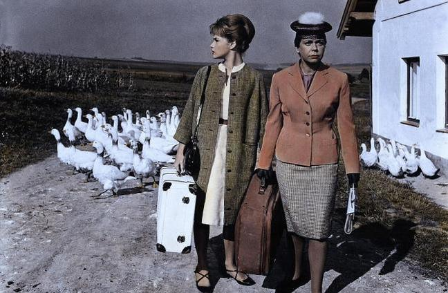 Zwei Frauen mit Koffern stehen vor einem Haus, während eine Schar Gänse vorbeizieht.