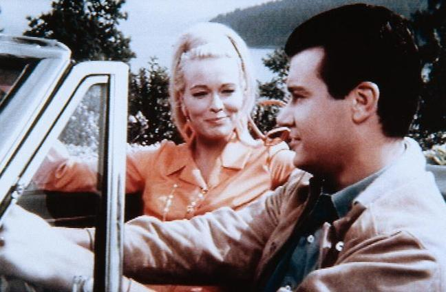 Ein Mann und eine Frau sitzen in einem Auto, im Hintergrund Bäume und Wasser.