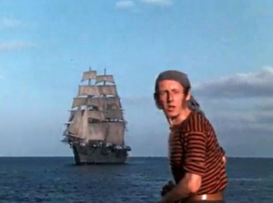 Ein Mann in einem gestreiften Hemd und Kopftuch blickt nach vorne, ein Schiff segelt auf dem Meer.