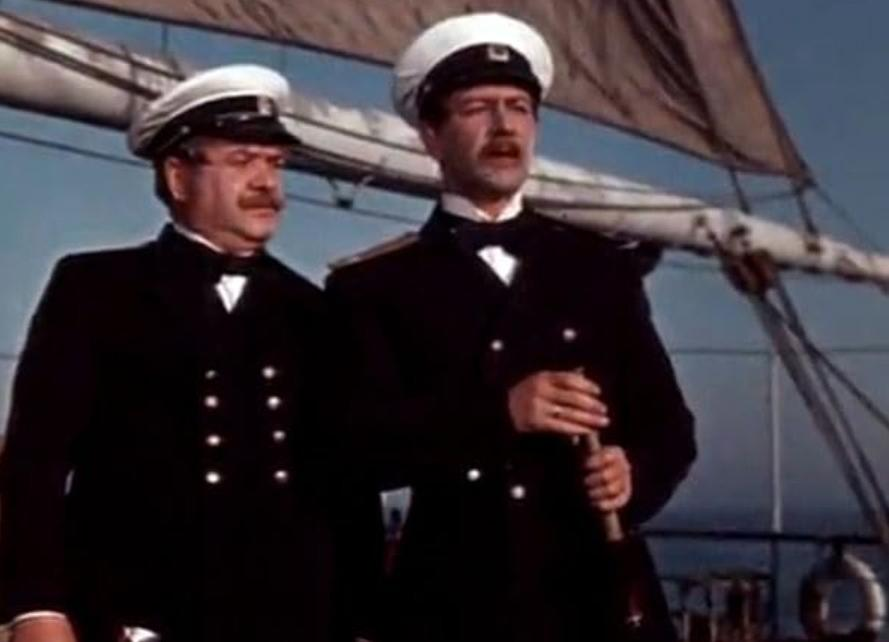 Zwei Männer in dunklen Uniformen und weißen Mützen stehen auf einem Segelschiff.