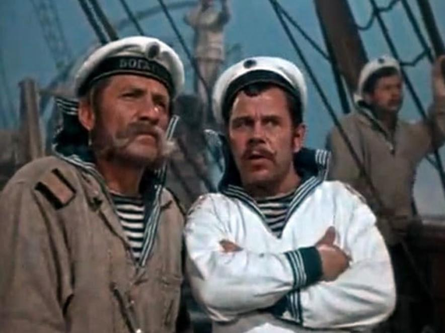 Zwei Seeleute in Matrosenuniformen stehen auf einem Deck. Einer trägt eine braune Jacke, der andere eine weiße.