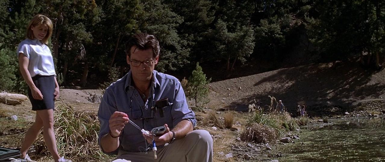 Mann mit Brille untersucht Gerät, Frau steht daneben, Wald im Hintergrund.