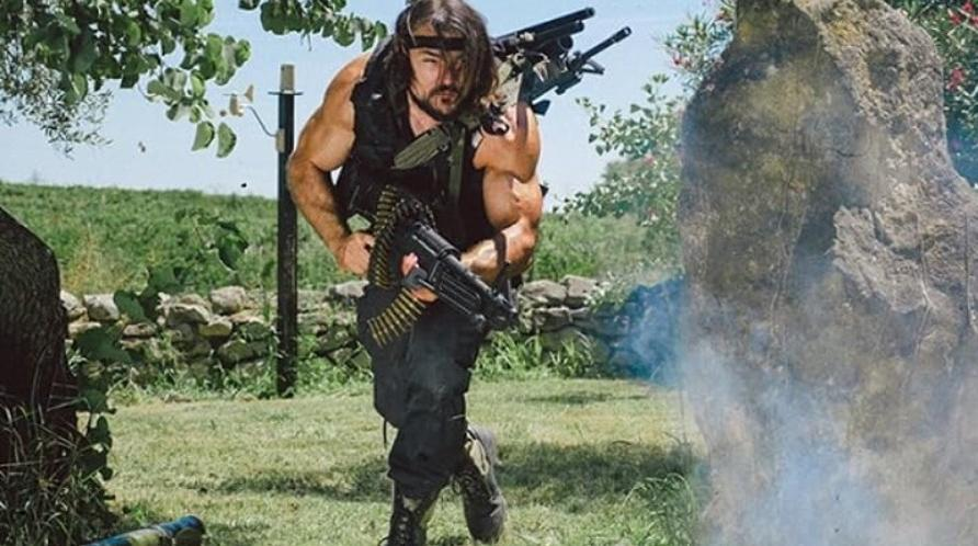 Ein Mann mit langen Haaren und Muskeln trägt Waffen und eine Munitionskette.