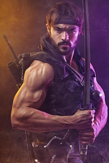 Ein Mann mit Bandana hält ein Schwert, trägt eine Weste und hat Waffen auf dem Rücken.
