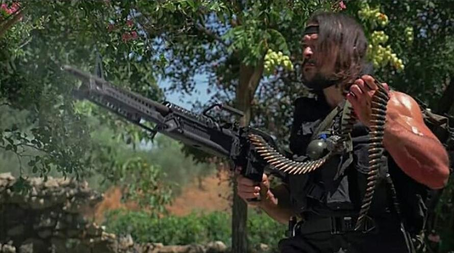 Ein Mann mit langen Haaren und einem Stirnband feuert ein Maschinengewehr ab.