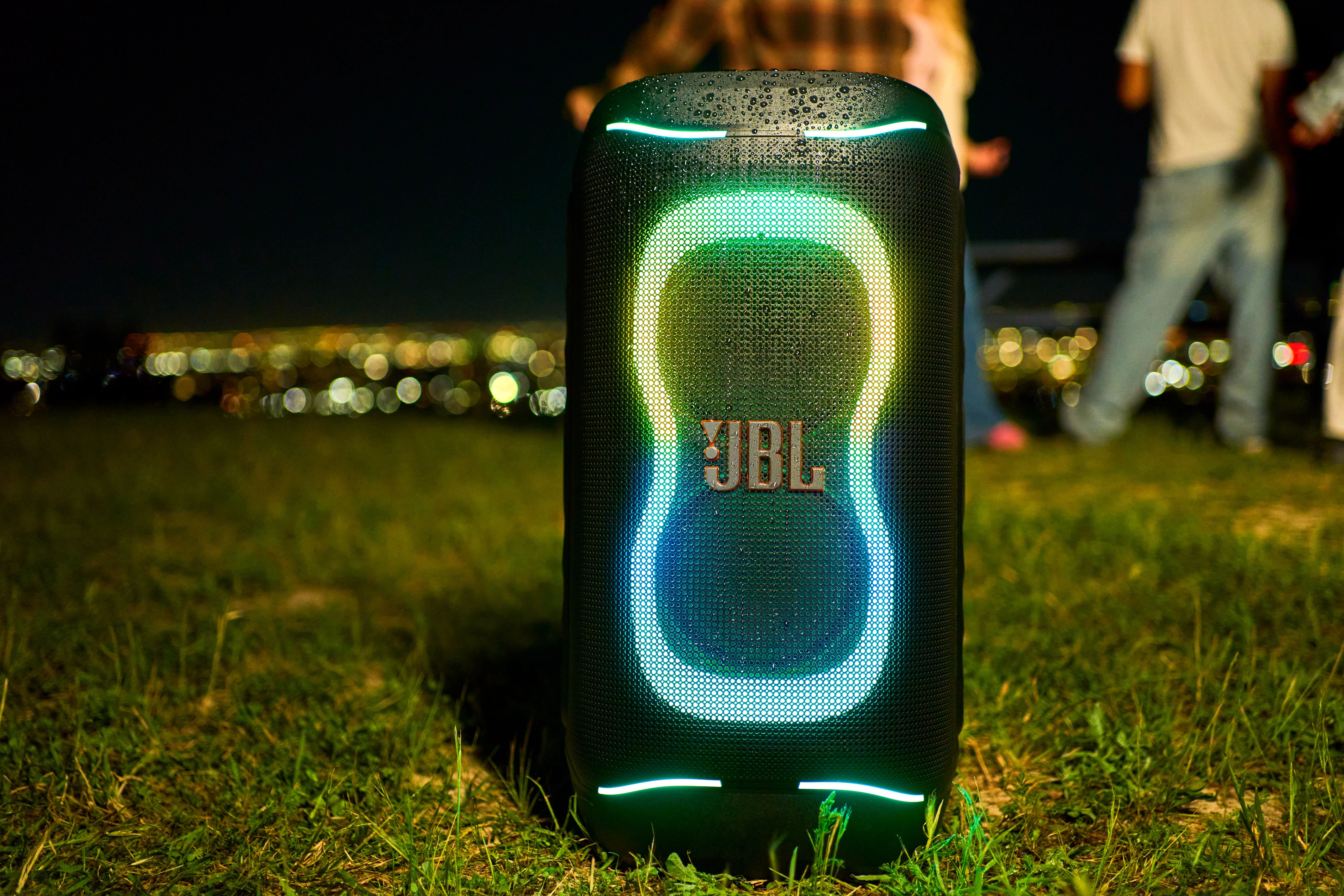 Gece çimenlerin üzerinde LED ışıklı bir JBL hoparlör. Arkada şehir ışıkları görünüyor.