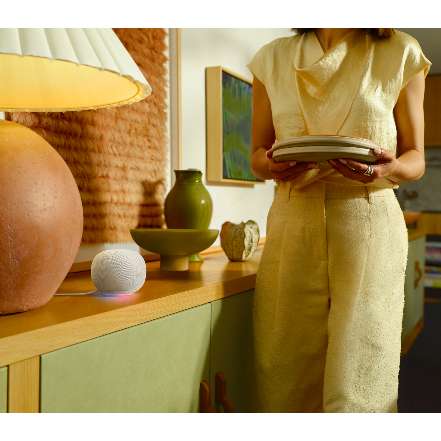 Een witte smart speaker op een houten plank met een vrouw die vaat vasthoudt.