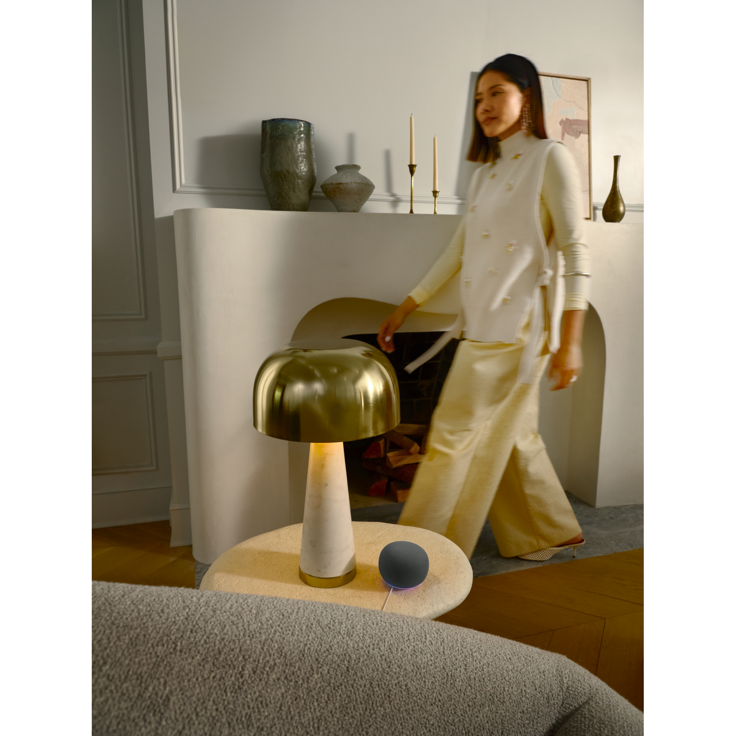 Een vrouw in een wit outfit loopt langs een open haard met een kleine speaker erop.